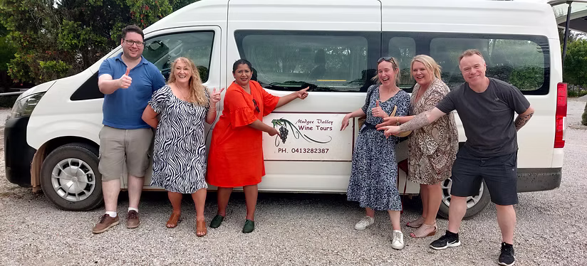 Group of six smiling people posing and pointing at a white van with 'Mudgee Valley Wine Tours' sign on its side.